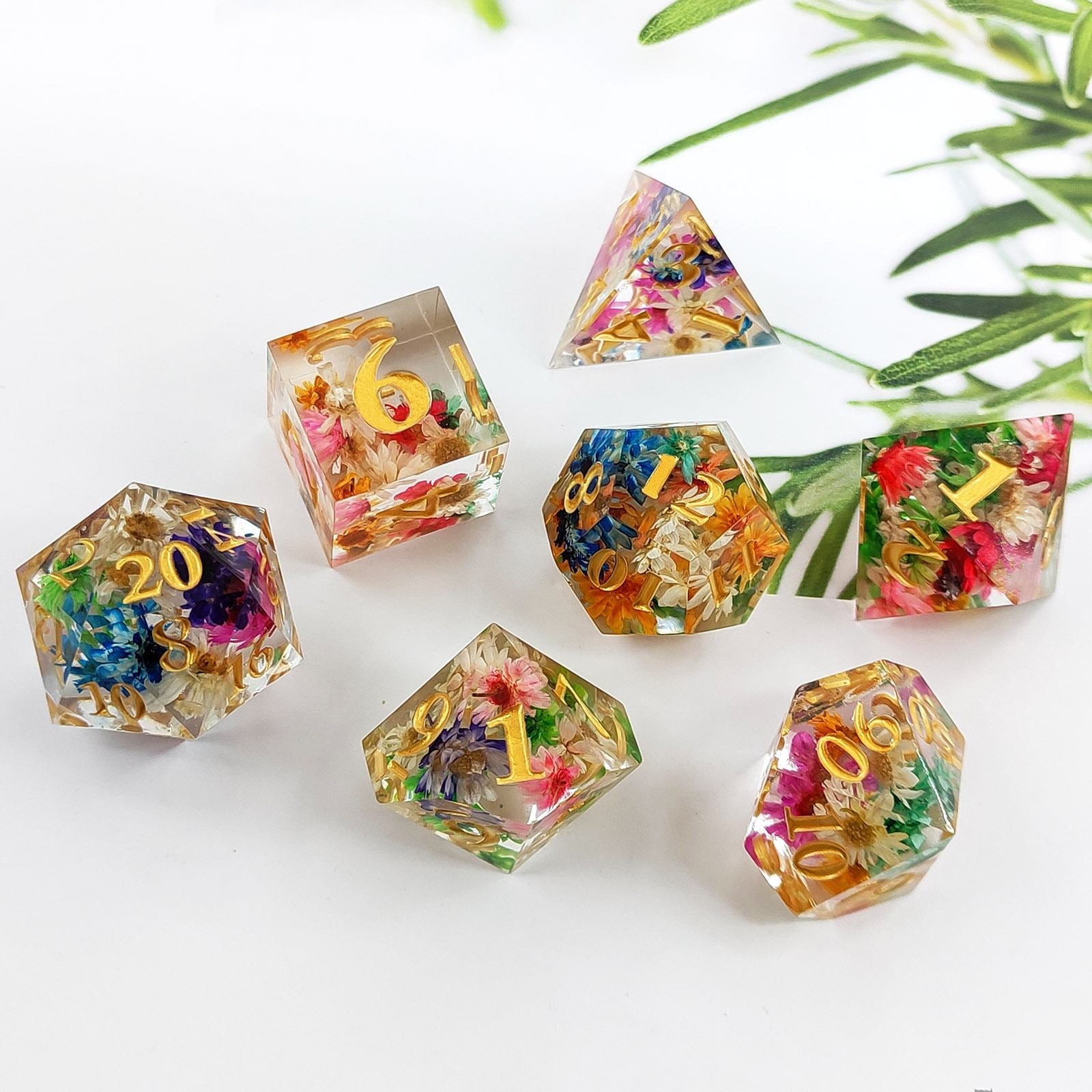 Set of colorful Sharp Edge Polyhedral resin dice with floral and letter embedded, on a white background with green leaves.