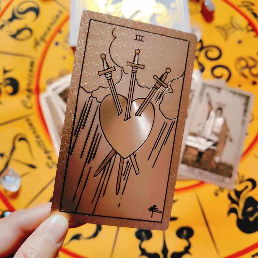 Rose Gold Holographic Tarot card with three swords piercing a heart held over a yellow background with tarot cards.